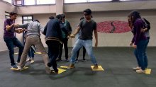 A group of teenagers doing an icebreaker exercise in which one person is blindfolded and the others are helping him jump across yellow tiles that are scattered across the floor